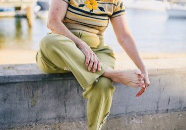 A woman sitting on a concrete ledge near the water, adjusting her shoe.